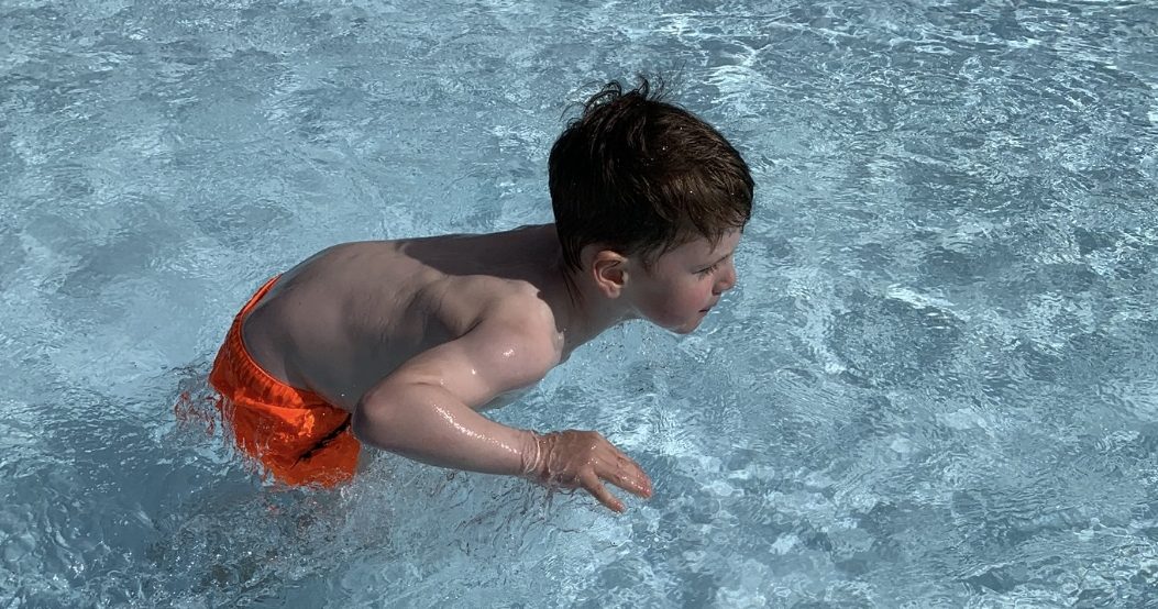 Ein Junge in orangefarbener Badehose watet durch einen blauen Pool.