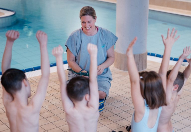 Eine fröhliche Schwimmlehrerin sitzt mit 4 Kindern im Kreis, die Kinder freuen sich und heben die Arme in die Luft.